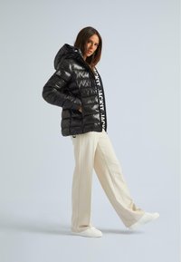 Giacca imbottita nera con una texture lucida, dotata di cappuccio e chiusura con zip, abbinata a pantaloni larghi color crema e sneakers bianche.