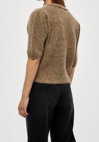 Žena v hnědém strukturálním krátkém svetru s krátkým rukávem a černých džínách s vysokým pasem, stojící bokem s viditelnými detaily na zádech a rukou.
