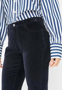 Vrouw in donkerblauwe corduroypantalon en een blauw-wit verticaal gestreept overhemd met lange mouwen, hand ontspannen langs haar zijde.