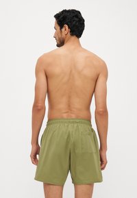 Shorts de bain vert olive en tissu léger, avec une taille élastique et une poche arrière unique. Texture lisse et longueur mi-cuisse.
