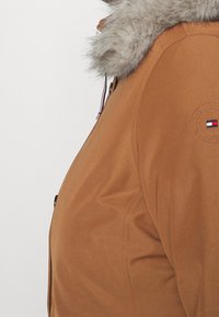 Chaqueta aislante marrón con un cuello de piel gris desmontable, cremallera lateral y un parche de logo circular con acentos en rojo, blanco y azul.