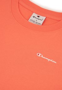 Koraalkleurig T-shirt van zachte stof, met een ronde halslijn en een wit "Champion" logo op de linkerkant van de borst. Maat S label zichtbaar.