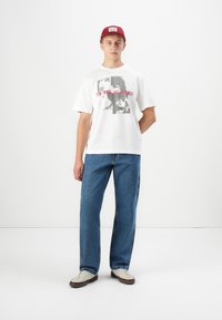 Ung mand iført en rød kappe, hvid Rolling Stones T-shirt med grafik, blå jeans og hvide sneakers, stående foran en helt hvid baggrund.