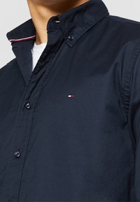 Man som bär en mörk marinblå skjorta med knappar och en liten röd, vit och blå logotyp på bröstet över en vit T-shirt.