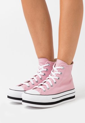 Jambes portant des baskets montantes roses à plateforme avec des lacets blancs et des semelles épaisses blanches comportant des rayures noires sur un fond blanc.