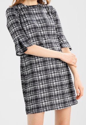 Žena v černobílých kostkovaných šatech po kolena s loketními rukávy stojí se založenýma rukama před sebou.