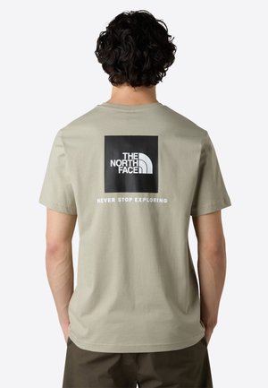 Mężczyzna z kręconymi włosami noszący beżowy t-shirt The North Face z czarnym logo i tekstem "Never Stop Exploring" na plecach.