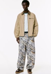 Uomo con una giacca beige, camicia bianca, pantaloni larghi a motivo astratto, cintura nera, scarpe marroni e collana in argento su sfondo bianco.