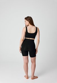 Woman standing barefoot facing away, wearing black crop top and high-waisted biker shorts against plain light background.