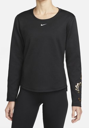 Femme portant un haut Nike noir à manches longues avec petit logo swoosh blanc sur la poitrine et motif floral doré sur la manche gauche.