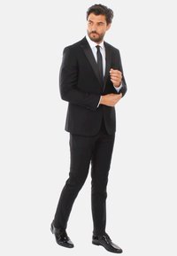 Tuxedo nero con rever in raso, camicia bianca e cravatta nera. Indossato con scarpe nere lucide. Silhouette slim fit, texture del tessuto liscia.