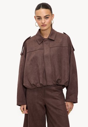 Vrouw in een oversized bruine suède jas met brede mouwen en bijpassende broek, geaccessoireerd met gouden oorringen en ringen, staande.
