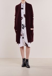 Cardigan in magenta a maglia con maniche lunghe e apertura frontale, abbinato a un vestito camicia bianco con motivi astratti neri e marroni e stivaletti neri alla caviglia.