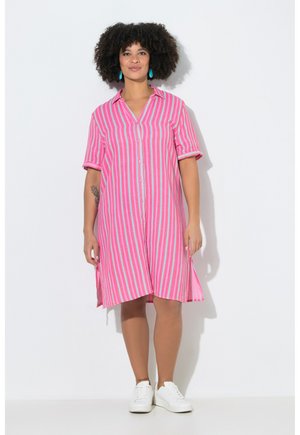 Kobieta stojąca, ubrana w sukienkę na guziki z pionowymi różowo-niebieskimi paskami, z krótkimi rękawami i białe trampki.