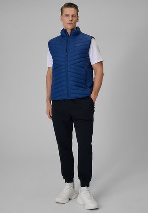 Homme portant un gilet matelassé bleu, une chemise blanche à manches courtes, un pantalon noir et des baskets blanches, debout devant un fond gris uni.