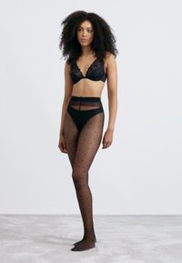 Soutien-gorge en dentelle noire, culotte taille haute noire et collants noirs transparents avec un petit motif à pois, présentés sur un modèle se tenant sur une surface claire.
