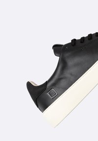 Sneaker in pelle nera con profilo basso, suola bianca spessa, punta rotonda e dettagli testurizzati. Presenta lacci neri e un logo semplice.