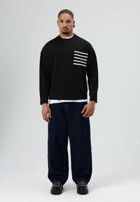 Zwart shirt met lange mouwen en een gestreepte zak, gecombineerd met losse donkere spijkerbroek en zwarte schoenen. Eenvoudig, casual design met een ontspannen pasvorm.