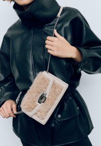 Osoba v černé kožené bundě drží béžovou crossbody tašku s texturou beránka a stříbrným řetízkem a sponou.