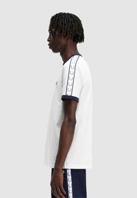 Camiseta de algodón blanca con cuello y puños de canalé en azul marino. Presenta un logo en el pecho y una franja decorativa con un patrón repetido a lo largo de las mangas.