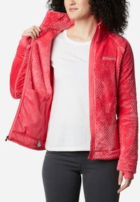 Giacca in pile di corallo con texture a rombi, chiusura con zip, tasca in rete interna e polsini elastici. Indossata sopra una camicia bianca e jeans grigi.
