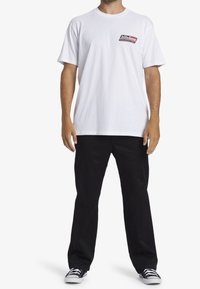 Bílá bavlněná tričko má na hrudi malé červené a černé grafické logo. Kombinováno s černými kalhotami a černobílými teniskami.