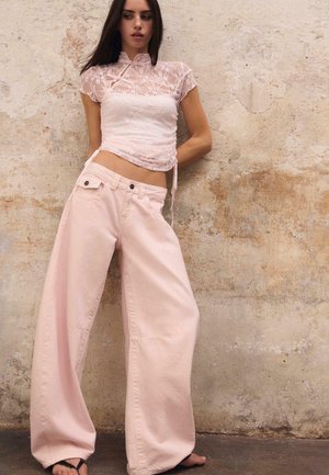 Femme debout contre un mur texturé, portant un haut en dentelle rose transparent et un pantalon large rose pâle avec des sandales noires, regardant vers sa gauche.