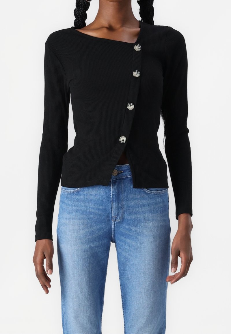 Femme portant un haut noir asymétrique à manches longues boutonné et un jean taille haute bleu clair, debout devant un fond blanc.