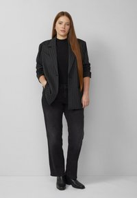 Blazer à fines rayures noires porté sur un haut noir ajusté, associé à un jean noir à jambes droites et des bottines noires à hauteur de la cheville. Cheveux longs et raides.