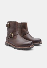 Stivali alla caviglia in pelle marrone con due cinture decorative, fibbie dorate e chiusura con zip. Suola robusta con finitura testurizzata.
