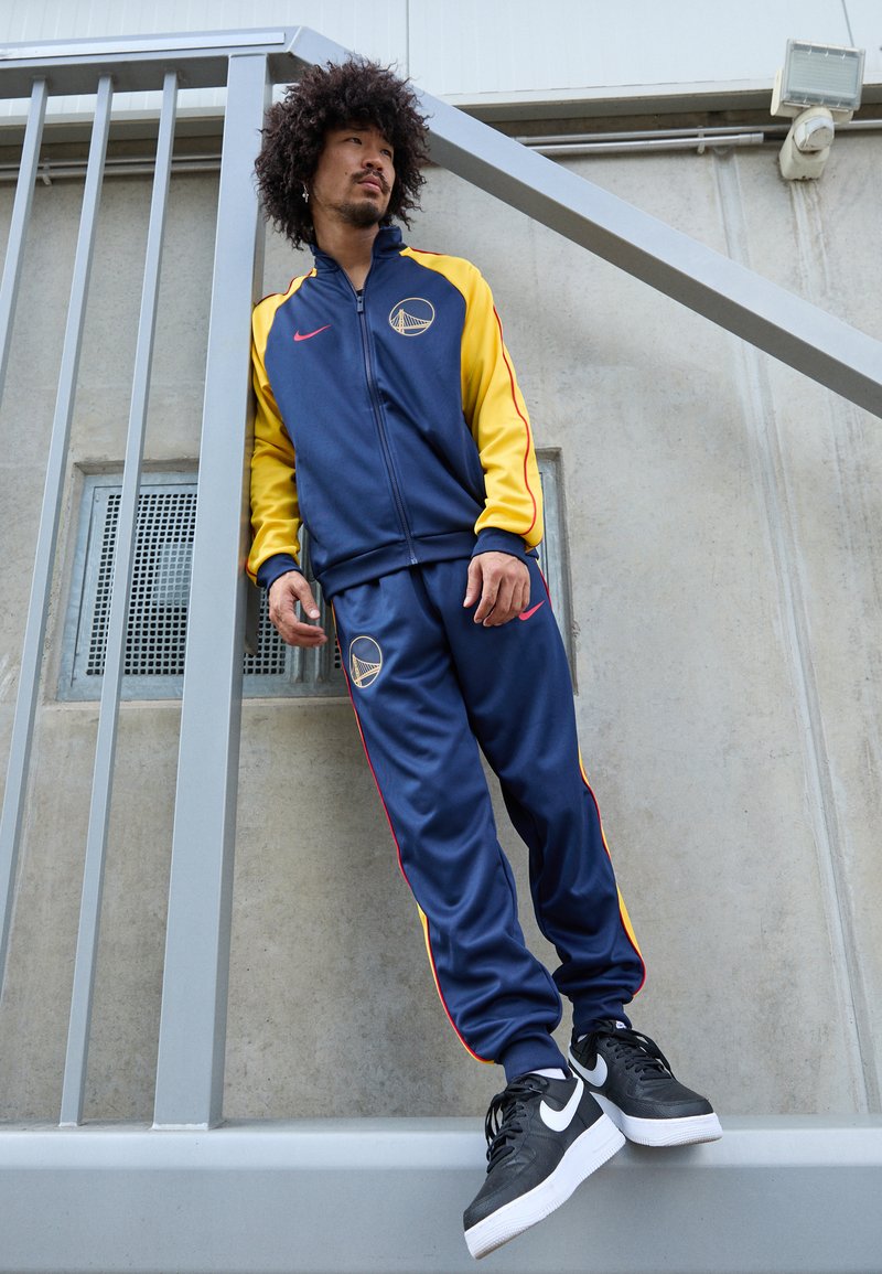 Tuta di colore blu navy con maniche gialle e dettagli rossi, caratterizzata da un logo sul petto. Sneakers nere con suole bianche e design testurizzato.