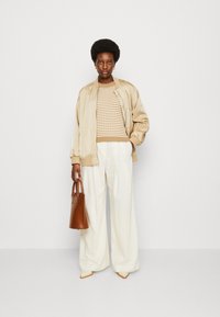 Giacca bomber beige sopra un maglione a righe, abbinata a pantaloni larghi bianchi a righe sottili. Tiene una borsa marrone e indossa scarpe beige a punta.