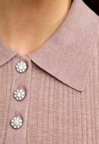 Close-up van een mauve geribbelde gebreide kraag met drie sprankelende strass-knopen vastgemaakt aan de nek en bovenste borst van een persoon.