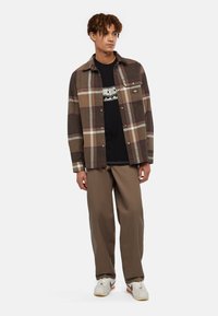 Camicia a quadri marrone con accenti beige, chiusura con bottoni, abbinata a una maglietta nera con grafica e pantaloni beige a gamba larga. Sneakers bianche completano il look.