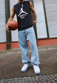 Persoon in een mouwloos shirt van de Brooklyn Nets, lichte spijkerbroek en witte sneakers, met een basketbal naast zich op een geplaveide stoep.