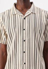 Short-sleeve shirt in cream fabric with black vertical stripes and dotted patterns, featuring a collared neck and black buttons.