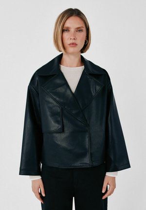 Femme aux cheveux blonds courts portant une veste en cuir noire oversize par-dessus une chemise blanche, se tenant devant un fond clair uni.