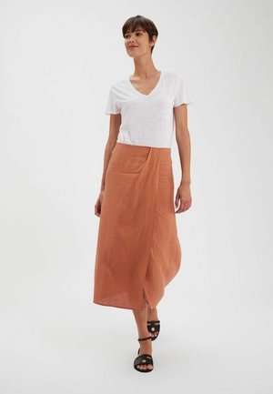 Jupe en lin rouille avec un design croisé à l'avant, associée à un t-shirt blanc uni à col en V et des sandales noires à lanières.
