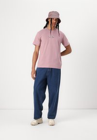 T-shirt de algodão rosa claro com um logótipo oval, combinado com jeans azul largos e um boné tipo balde rosa suave. Os sapatos são de uma tonalidade bege clara com detalhes texturizados.