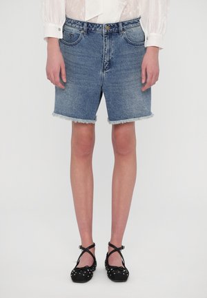 Personne portant un short en jean à ourlet effiloché, un chemisier blanc à manches longues et des ballerines noires cloutées avec des brides à la cheville.