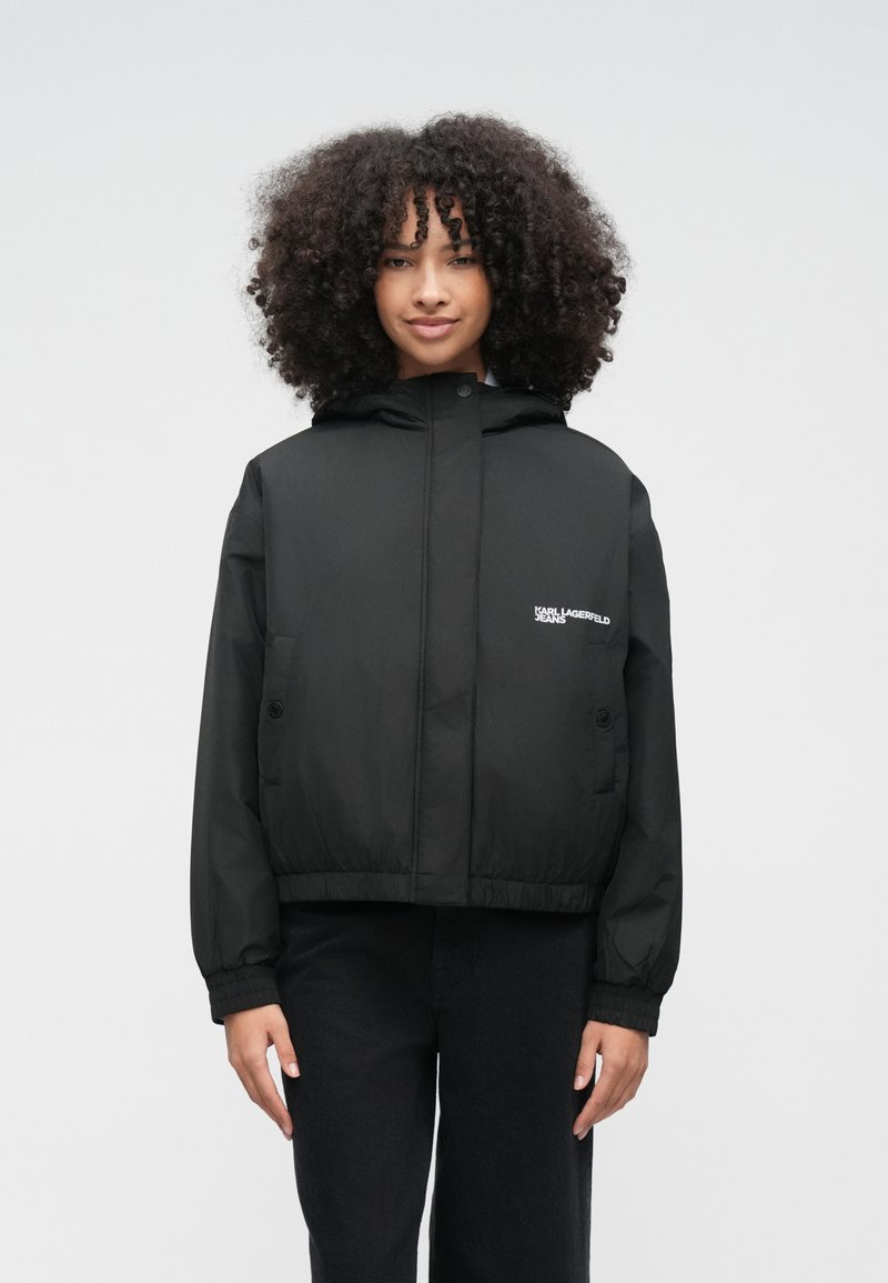 Mladá žena s kučeravými vlasmi, oblečená v čiernej kapucňovej prešívanej bunde a čiernych nohaviciach, stojaca pred jednoduchým bielym pozadím.