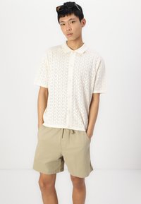 Short-sleeve, cream-colored polo shirt with a textured chevron pattern, paired with beige shorts, featuring an elastic waistband.
