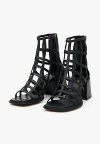 Black heeled sandals with open toes, crisscross straps, and block heels. Made of smooth synthetic material with a matte finish.