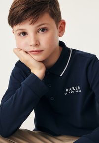 Tumši zilas krāsas garām piedurknēm polo krekls ar baltu apkakles apdari, ar pogu aizdari un uzkrāsotu tekstu "BAKER BY TED BAKER" uz krūškurvja.