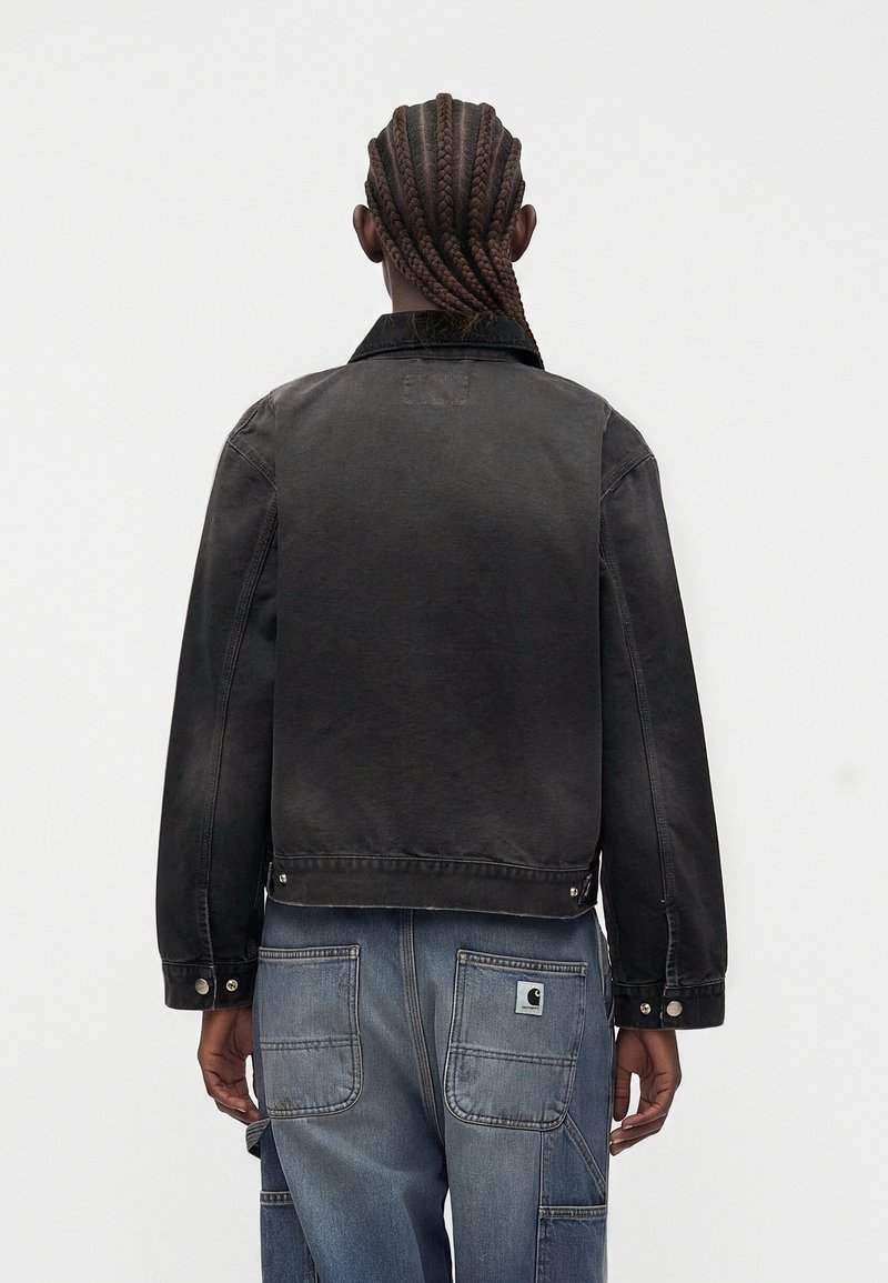 Personne aux cheveux tressés portant une veste en jean noire et un jean bleu avec des poches appliquées, vue de dos sur un fond uni.