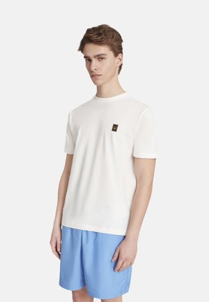 Giovane uomo in piedi, indossa una maglietta bianca semplice con una piccola toppa sul petto e pantaloncini larghi azzurri chiari su sfondo bianco.