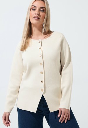 Kvinde med langt blondt hår iført en cremefarvet knap-op sweater og mørkeblå jeans mod en ensfarvet lys baggrund.
