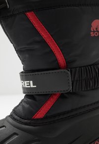 Czarne wodoodporne buty śniegowe z czerwoną akcentującą paskiem, regulowanym syntetycznym paskiem z logo "SOREL" oraz teksturowaną gumową podeszwą.