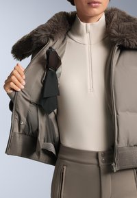 Barna steppelt dzseki szőrmés gallérral, kigombolatlanul, alatt világos krémszínű aláöltözet látható. Egy fekete anyag és egy napszemüveg is látható benne.