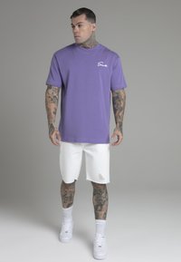 Lilla crew neck t-shirt i bomuld med hvidt logo; parret med slidte hvide shorts og hvide sneakers. Mønstret hudtatoveringer synlige.